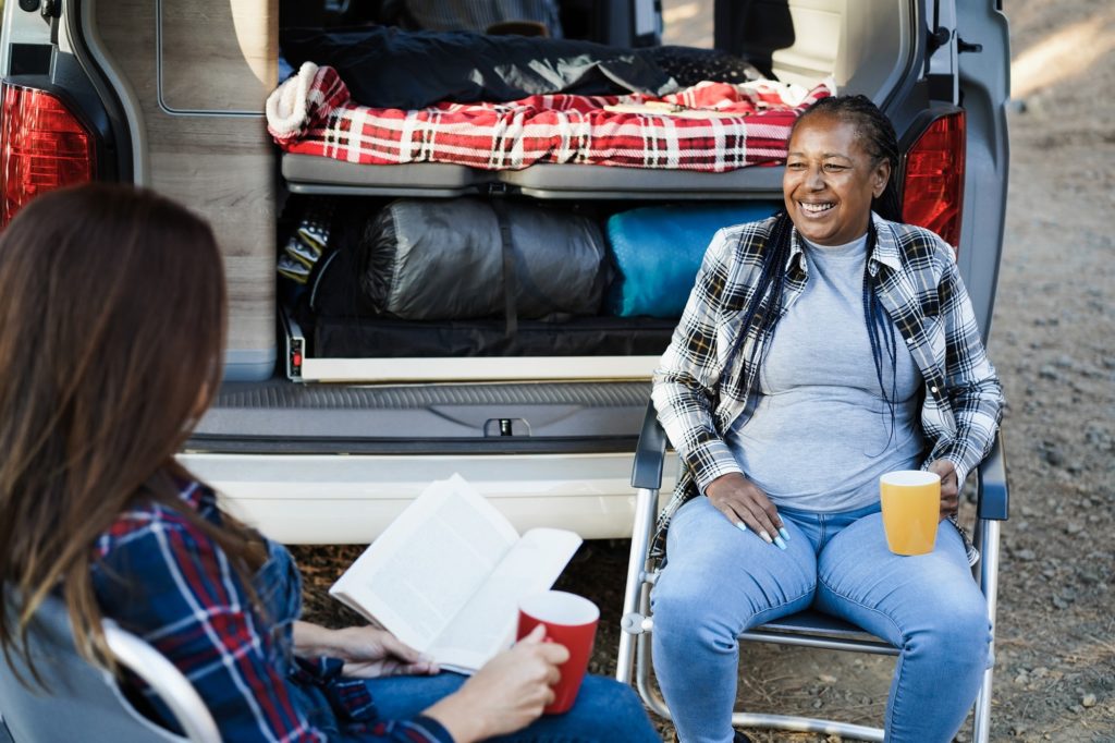 Multiracial women friends having fun camping with camper van while reading and drink coffee